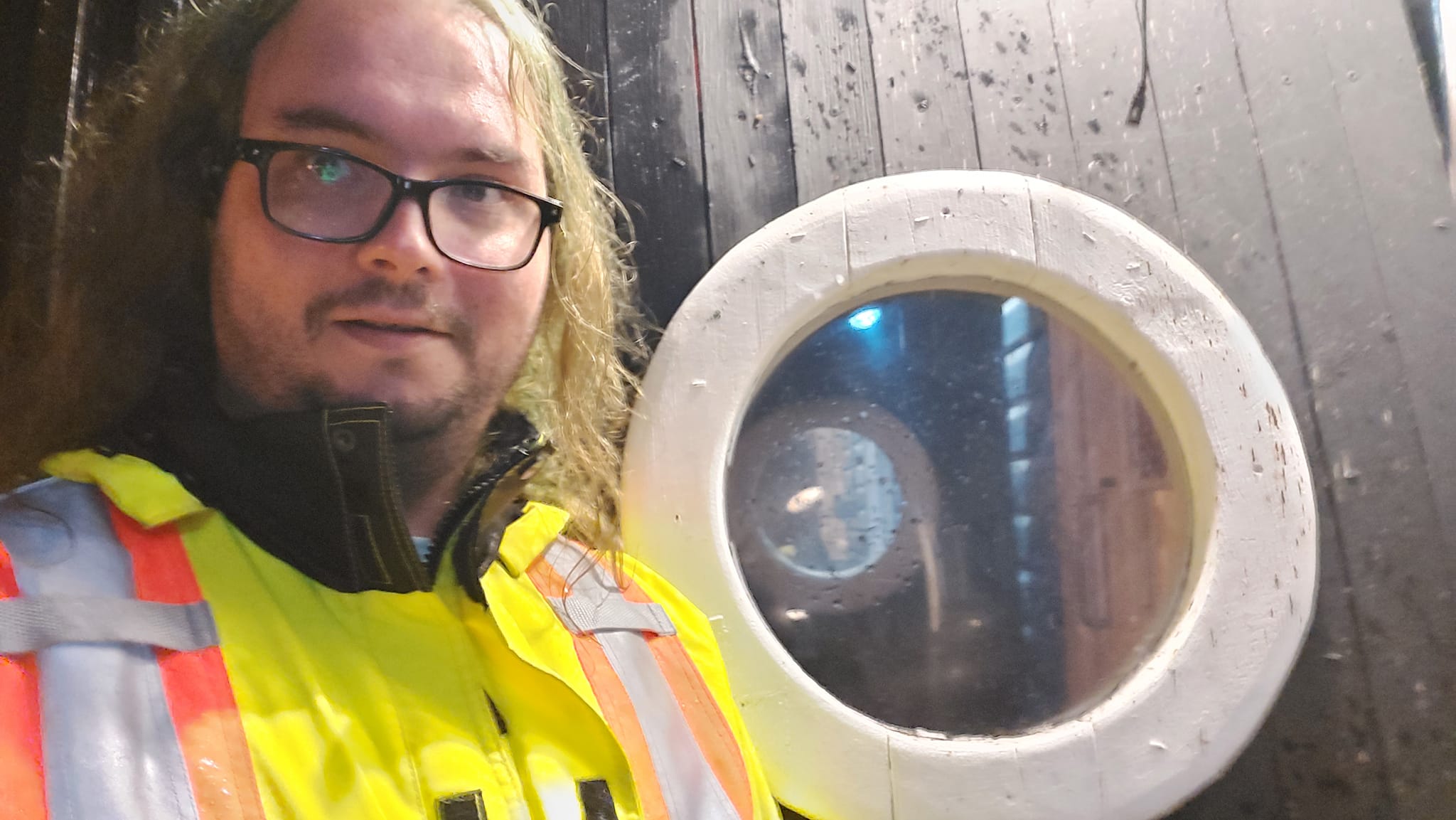 A man with long green hair and no beard wears a high visibility raincoat in front of a port hole like window